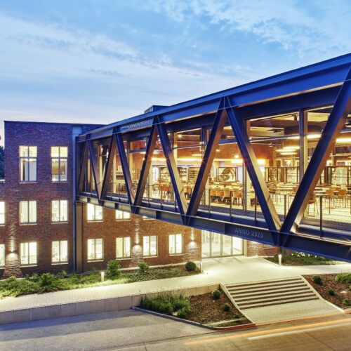Verwaltungsgebäude von Big Dutchman in Calveslage mit verglaster Verbindungsbrücke, entworfen von Geising + Böker Architekten – moderne Büroarchitektur bei Abendlicht.
